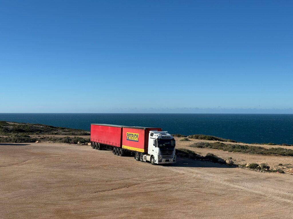 A white truck with a red trailer travels on a coastal road next to the ocean.